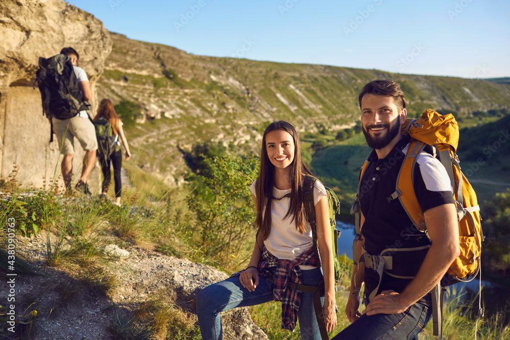 Portrait of a couple of young hikers with backpacks on a background of rocky mountains. Guy and girl are resting and smiling at the camera. Concept of hiking, tourism, nature, people and lifestyle.