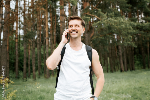 Happy man talking on a cell phone while walking in the woods. Athletic traveler with backpack smiling and holding phone, pine forest outdoors background. Handsome young man lifestyle