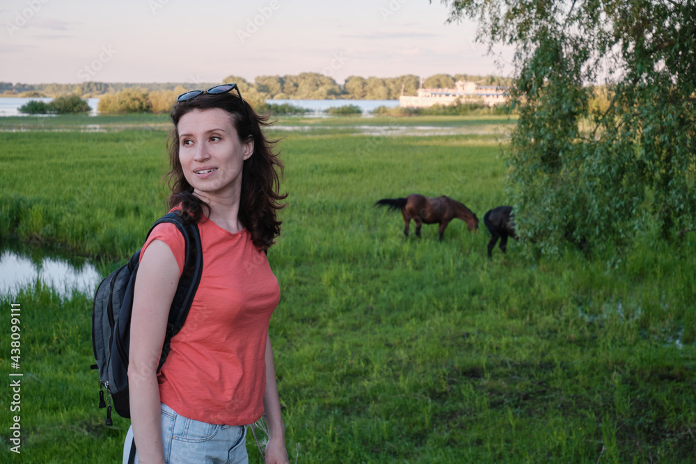 Girl with a backpack, green meadow, two horses in the background.
