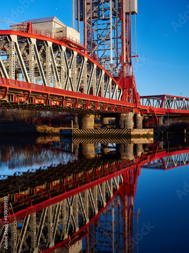 The once-famous, now de-commissioned Newport Bridge on the River Tees. The red bridge contrasts with the blue sky, both reflected in glassy river.