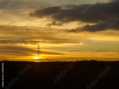 An electricity pylon, back-lit and silhouetted by the setting sun with an intricate, layered cloudscape in a huge golden sky.