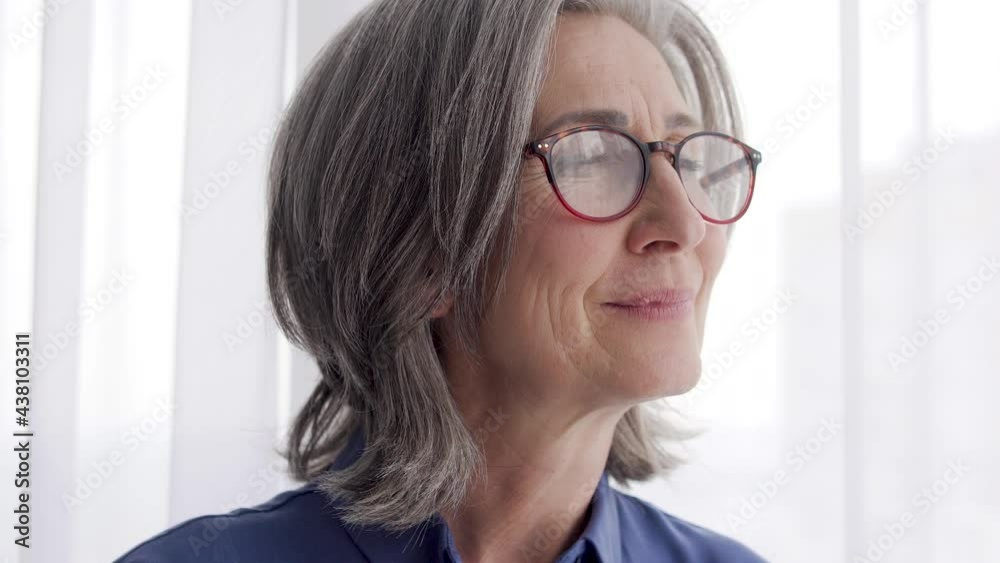 Positive middle aged woman in eyeglasses smiling enjoying view from windows Positive middle aged woman in eyeglasses smiling enjoying view from windows