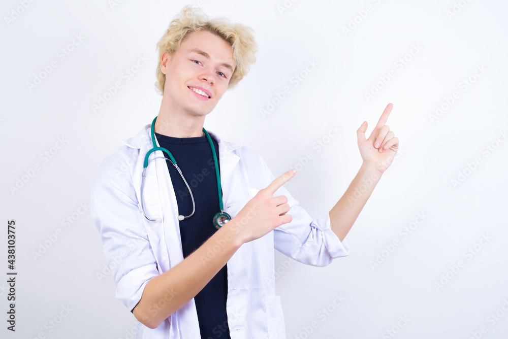 young handsome Caucasian doctor man standing against white background with positive expression, indicates with fore finger at blank copy space for your promotional text or advertisement. young handsome Caucasian doctor man standing against white background with positive expression, indicates with fore finger at blank copy space for your promotional text or advertisement.