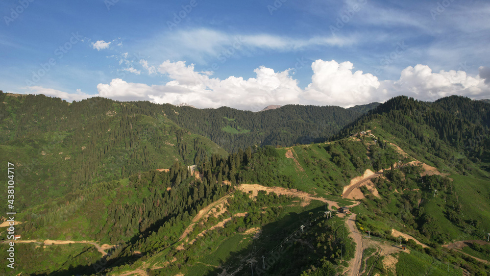 Green hills and mountains with white clouds. A prominent road, pillars, gondola road and wooden houses. The mountains are covered with coniferous forest. Blue sky. Oi-Qaragai Mountain resort Almaty