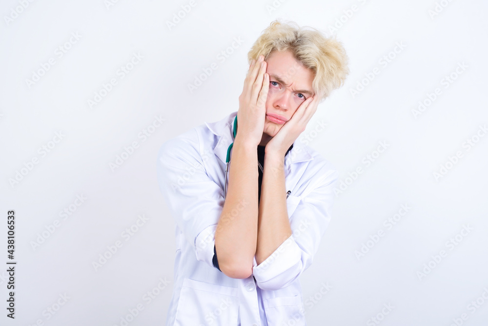 young handsome Caucasian doctor man standing against white background holding head in hands with unhappy expression watching sad movie about animals and trying not to cry. young handsome Caucasian doctor man standing against white background holding head in hands with unhappy expression watching sad movie about animals and trying not to cry.