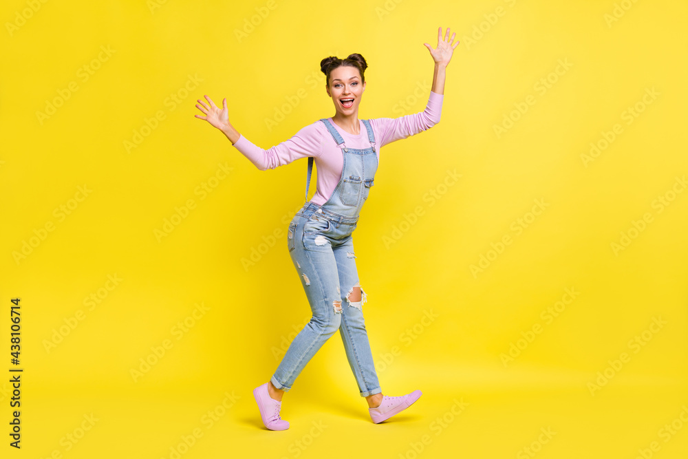 Full length photo of charming shiny young woman wear denim overall dancing rising arms hands isolated yellow color background