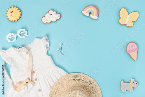 Stylish summer set of child clothes. White dress, the straw hat, sunglasses and funny gingerbread on blue background. Fashion girl lookbook concept. Top view Flat lay Copy space