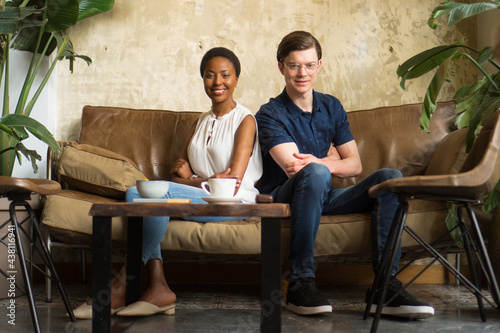 A young man and a woman are posing for the photo after the successful business meeting. They sit on a brown leather sofa in front of a beige wall with big houseplants on each side.