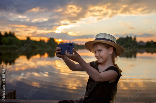 Pretty preteen girl in summer straw hat taking selfie for cellphone at beautiful sunset on lake, lifestyle, local travel concept, young blogger