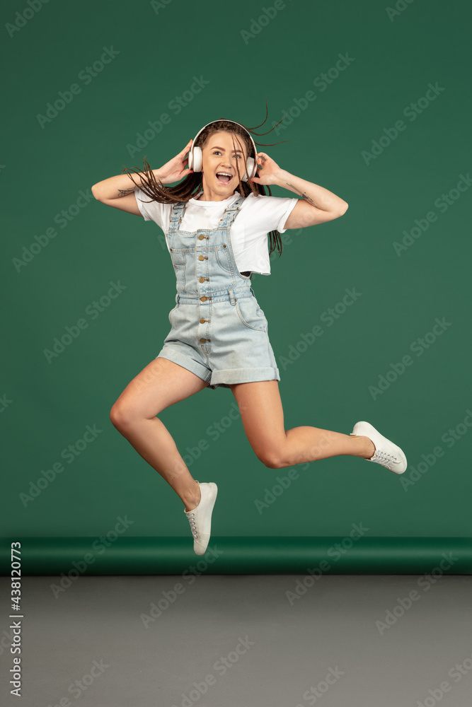 Caucasian woman with headphones jumping over green studio background with copyspace Caucasian woman with headphones jumping over green studio background with copyspace