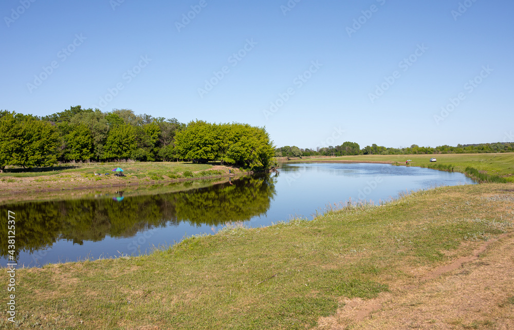 Several anglers are fishing on a clear, sunny summer day on a small, picturesque river.