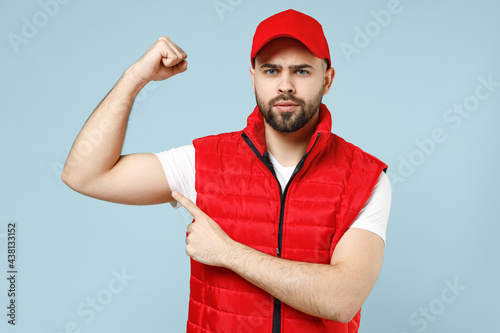 Strong sporty delivery guy employee man in red cap white T-shirt vest uniform workwear work as dealer courier showing biceps muscles on hand isolated on pastel blue background studio. Service concept.