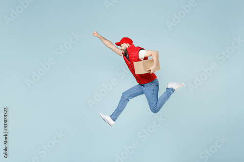 Full size body length side view delivery guy employee man in red cap white T-shirt vest uniform work dealer courier jump run go walk isolated on pastel blue color background studio. Service concept photo