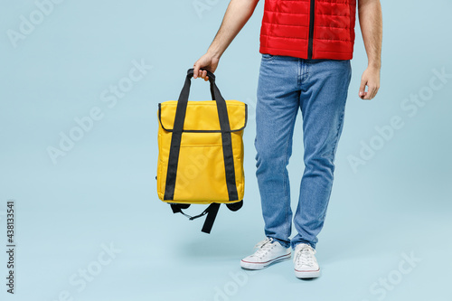 Delivery guy employee man in red vest uniform jeans on legs workwear work dealer courier hold in arm yellow thermal food bag backpack isolated on pastel blue color background studio Service concept photo