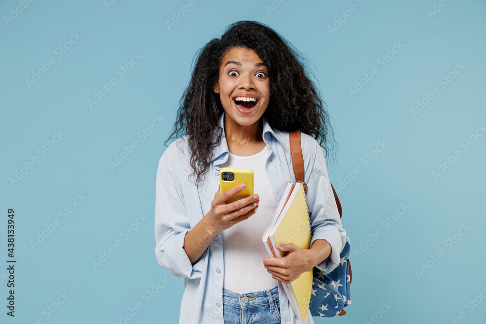 Young amazed african american girl teen student in denim clothes backpack hold books use mobile cell phone chat online isolated on blue background Education in high school university college concept