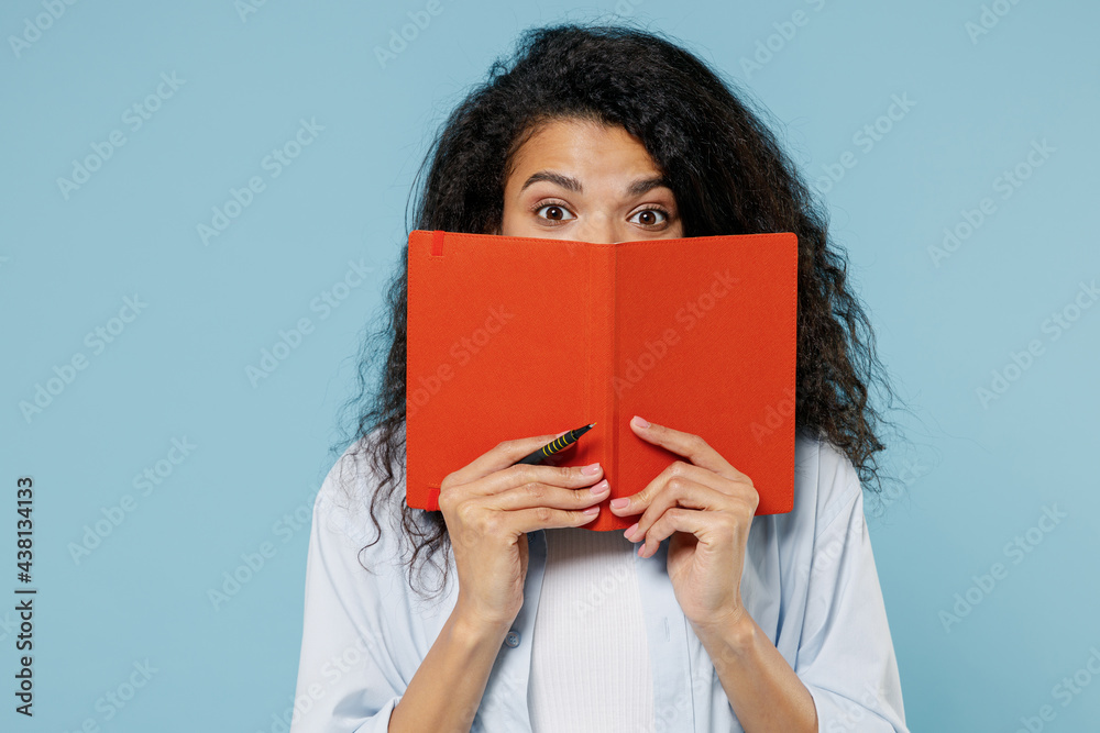 Young shocked puzzled nerd african american girl teen student in denim clothes cover mouth hiding with book notebook isolated on blue background Education in high school university college concept Young shocked puzzled nerd african american girl teen student in denim clothes cover mouth hiding with book notebook isolated on blue background Education in high school university college concept