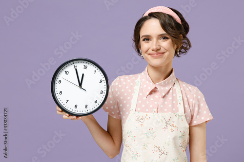 Young smiling happy cheerful fun housewife housekeeper chef cook baker woman wear pink apron hold clock look camera isolated on pastel violet background studio portrait Cooking food process concept.