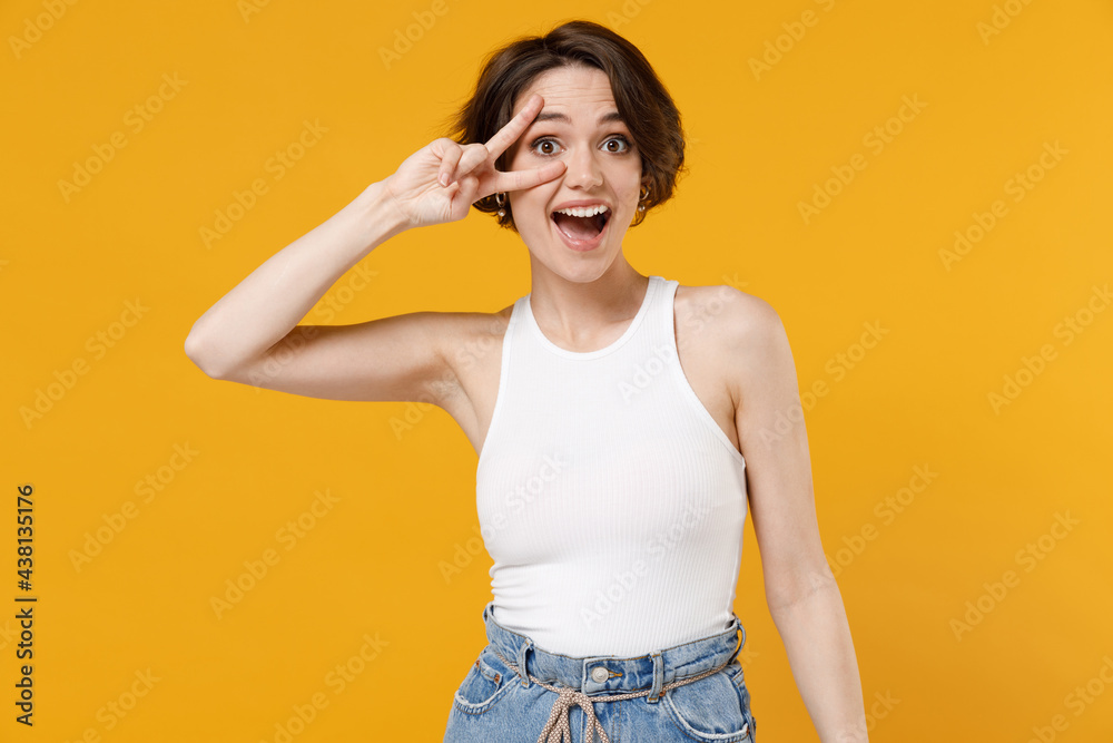 Young friendly smiling happy woman 20s with bob haircut in white tank top shirt cover eye with victory v-sign gesture look camera isolated on yellow color background studio People lifestyle concept Young friendly smiling happy woman 20s with bob haircut in white tank top shirt cover eye with victory v-sign gesture look camera isolated on yellow color background studio People lifestyle concept