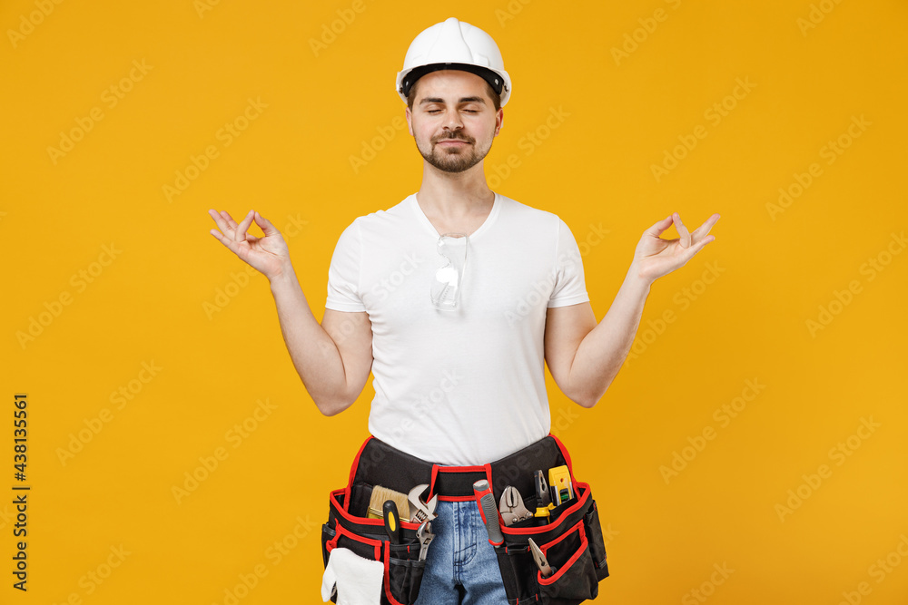 Young employee handyman man in protective helmet hardhat hold arm in yoga om gesture meditate realx isolated on yellow background Instruments accessories renovation apartment room Repair home concept.