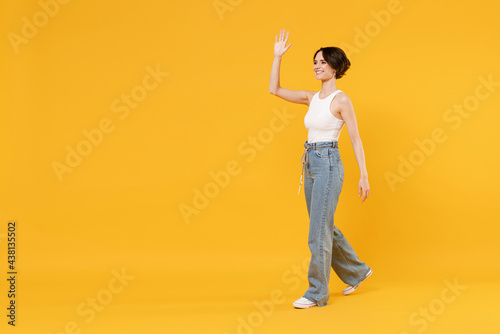 Full length side view young smiling happy woman 20s with bob haircut wearing white tank top shirt walking going look aside waving hand isolated on yellow background studio People lifestyle concept