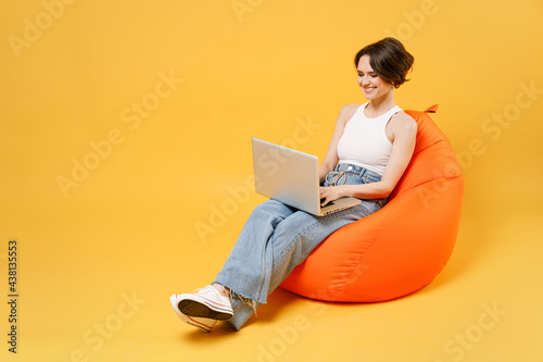 Young smiling woman 20s with bob haircut wearing white tank top shirt using laptop pc computer chat online browsing surfing internet sit in orange bag chair hold face isolated on yellow background