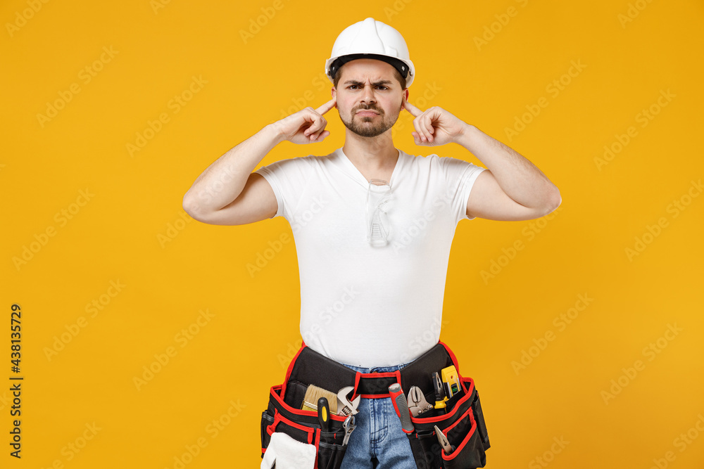 Young employee handyman man in protective helmet hardhat cover ears hand fingers do not want listen isolated on yellow background Instruments accessories renovation apartment room Repair home concept Young employee handyman man in protective helmet hardhat cover ears hand fingers do not want listen isolated on yellow background Instruments accessories renovation apartment room Repair home concept