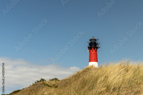 a red and white striped lighthouse stands on a dune, in the foreground you can see beach grass growing. a lighthouse stands against a bright blue sky with light white veil clouds.