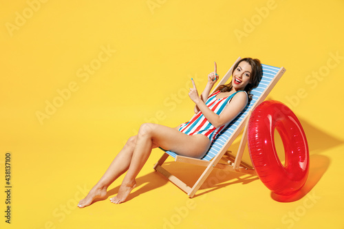 Full body length happy young sexy woman wear striped red blue one-piece swimsuit sit on wooden chair isolated on vivid yellow color wall background studio. Summer hotel pool sea rest sun tan concept.