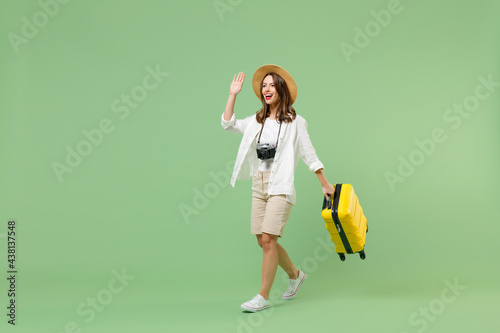 Full length traveler tourist woman in casual clothes straw hat hold yellow suitcase walk waving hand isolated on pastel green background. Passenger travel abroad weekends. Air flight journey concept.