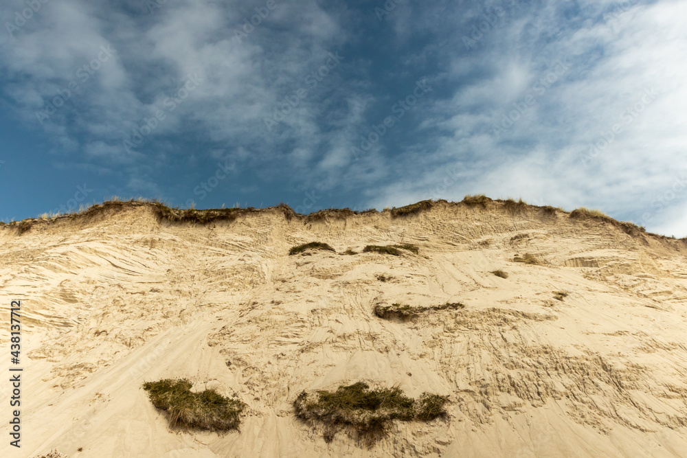 a sand dune with lots of grass overgrown in front of a bright blue sky with fine white veil clouds. 