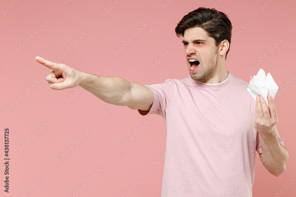Side view sick ill angry allergic man has watery red eyes runny nose suffer from allergy symptoms hold paper napkin scream pointing hand finger aside isolated on pastel pink color background studio. Side view sick ill angry allergic man has watery red eyes runny nose suffer from allergy symptoms hold paper napkin scream pointing hand finger aside isolated on pastel pink color background studio.