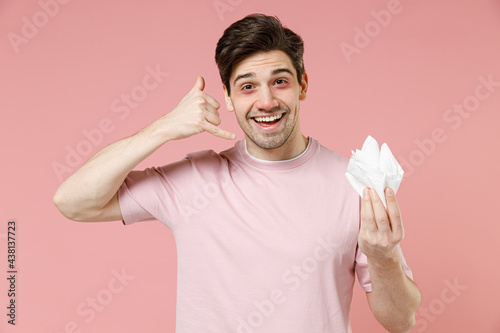 Sick ill allergic cheery man wear rozy t-shirt has red eyes runny nose suffer from allergy hold paper napkin doing phone gesture like says call me back isolated on pastel pink color background studio photo