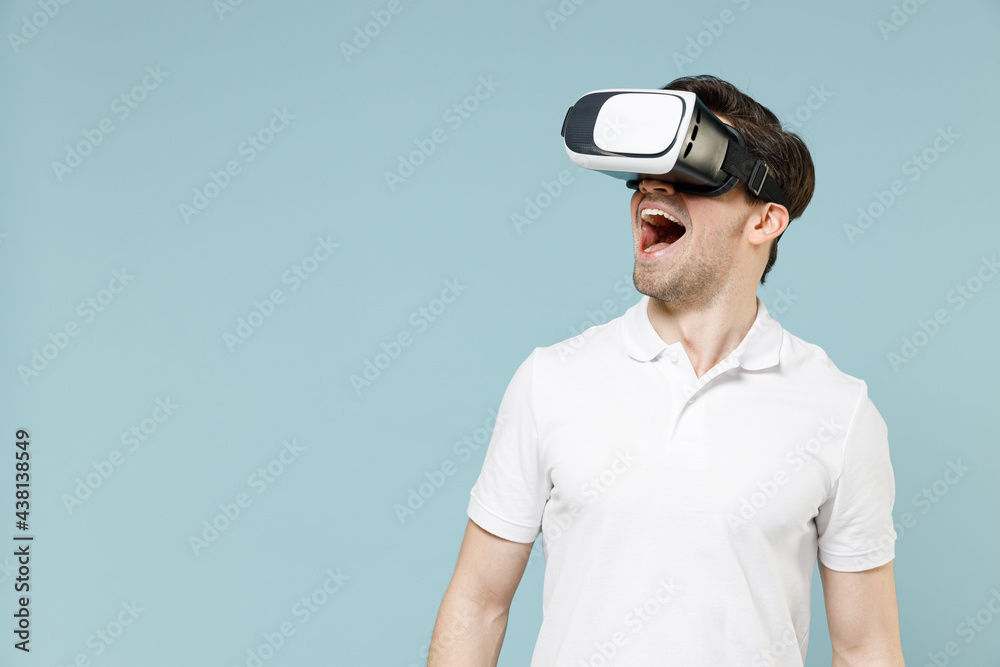 Young amazed surprised shocked man 20s in white casual basic t-shirt play pc game watching in vr headset pc gadget isolated on pastel blue color background studio portrait People lifestyle concept. Young amazed surprised shocked man 20s in white casual basic t-shirt play pc game watching in vr headset pc gadget isolated on pastel blue color background studio portrait People lifestyle concept.