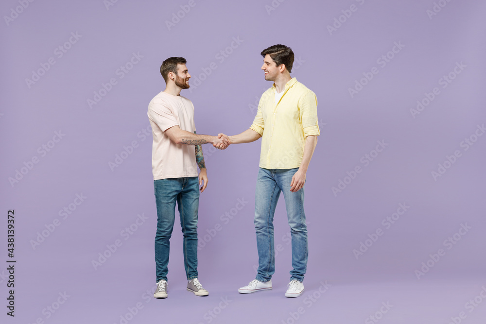 Full length two young smiling happy men friends together in casual t-shirt meeting together greet hold hands folded handshake gesture n isolated on purple background studio People lifestyle concept Full length two young smiling happy men friends together in casual t-shirt meeting together greet hold hands folded handshake gesture n isolated on purple background studio People lifestyle concept