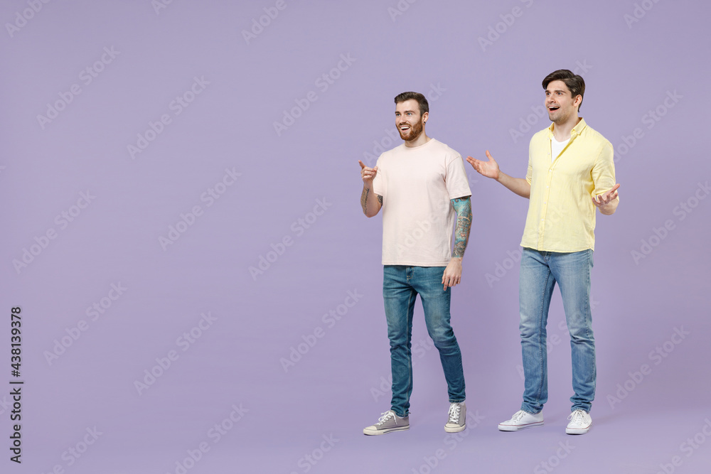 Full length shocked excited two young men friends together in casual t-shirt walk go point finger aside spread arms isolated on purple background studio portrait People friendship lifestyle concept
