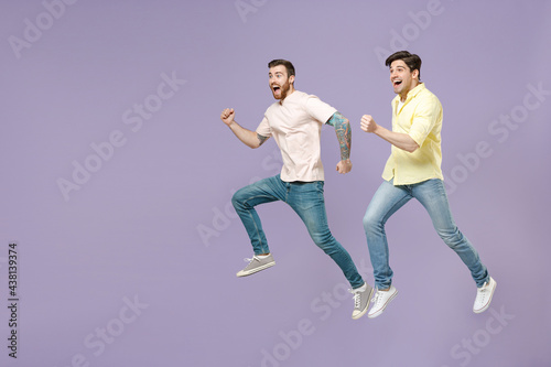 Side view full length two young overjoyed smiling men friends together in casual t-shirt looking to each other hurrying up run jump high isolated on purple background studio People lifestyle concept