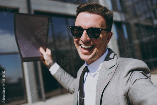 Close up young overjoyed employee business man in grey suit walk go near office glass wall building outdoors in downtown city center doing selfie shot on mobile phone show folder for papers documents © ViDi Studio