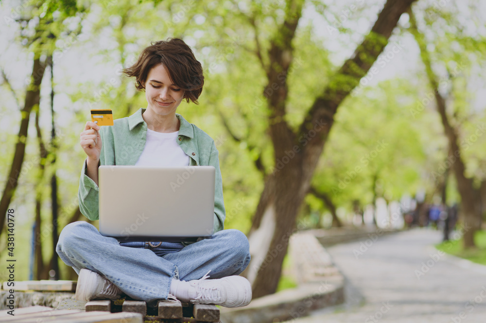 Full length young freelancer woman in green jacket jeans sit on bench in park outdoors use laptop pc computer credit bank card do online shopping order booking delivery People urban lifestyle concept. Full length young freelancer woman in green jacket jeans sit on bench in park outdoors use laptop pc computer credit bank card do online shopping order booking delivery People urban lifestyle concept.