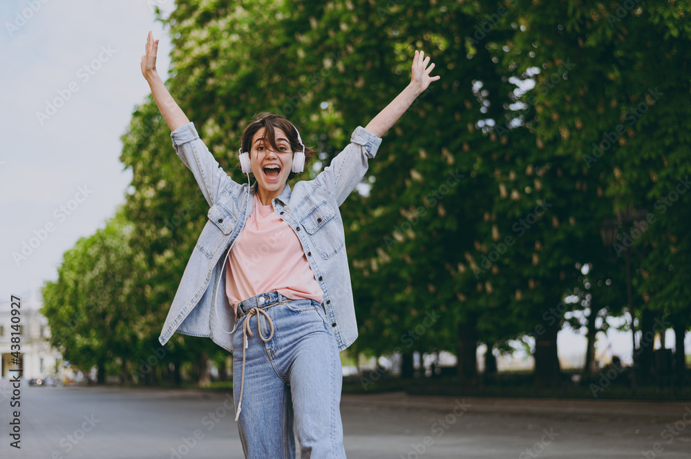 Young overjoyed happy student caucasian woman 20s wearing jeans clothes headphones listen to music walking strolling down green park alley outdoors with raised up hands People urban lifestyle concept Young overjoyed happy student caucasian woman 20s wearing jeans clothes headphones listen to music walking strolling down green park alley outdoors with raised up hands People urban lifestyle concept
