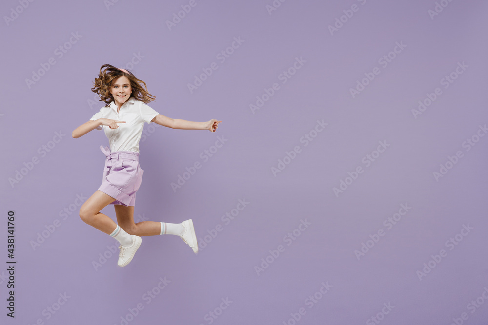 Full length little kid girl 12-13 years old in white shirt jumping high point index finger aside on workspace area mock up isolated on purple color background. Childhood children lifestyle concept