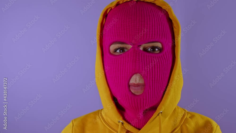 Close up of unrecognizable woman in pink balaclava shows her tongue on purple background. Unknown female in mask sticks out tongue and hides it, looking at camera