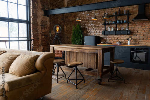 Spacious industrial kitchen and living room with big windows, brown sofa and black cabinets