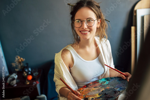 Indoor portrait of a pretty artist female sitting next to the easel with canvas painting something in her art studio. A young woman painter in transparent spectacles draws in the workshop.