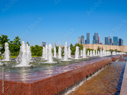 Jets of fountains on a sunny day against the backdrop of modern skyscrapers and a cloudless blue sky. Recreation area in Victory Park on Poklonnaya Hill in summer.