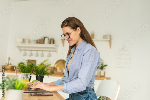 Attractive businesswoman studying online, using laptop software, web surfing information or shopping in internet store. Smiling young woman using laptop in the kitchen at home. Working from home in