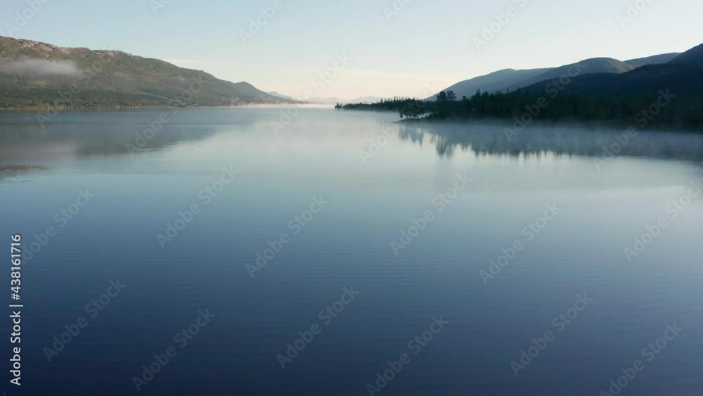 A low flight above the mirrorlike surface of a Nedre Fiplingvatnet lake. Morning mist is dissipating, revealing not yet lighted up black hills on the right. Dense pine forest is visible on the left. A low flight above the mirrorlike surface of a Nedre Fiplingvatnet lake. Morning mist is dissipating, revealing not yet lighted up black hills on the right. Dense pine forest is visible on the left.