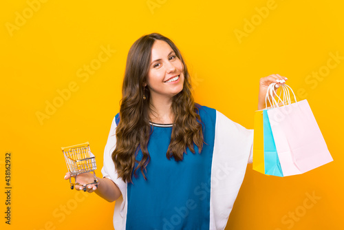 Charming young woman standing over yellow background and holding shopping bags and shopping trolley