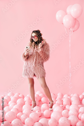 Full length portrait of glamour young woman typing on mobile smartphone over pink balloons background photo