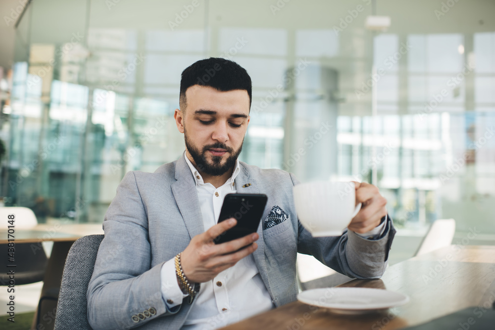 Serious businessman browsing smartphone and enjoying cup of hot drink Serious businessman browsing smartphone and enjoying cup of hot drink