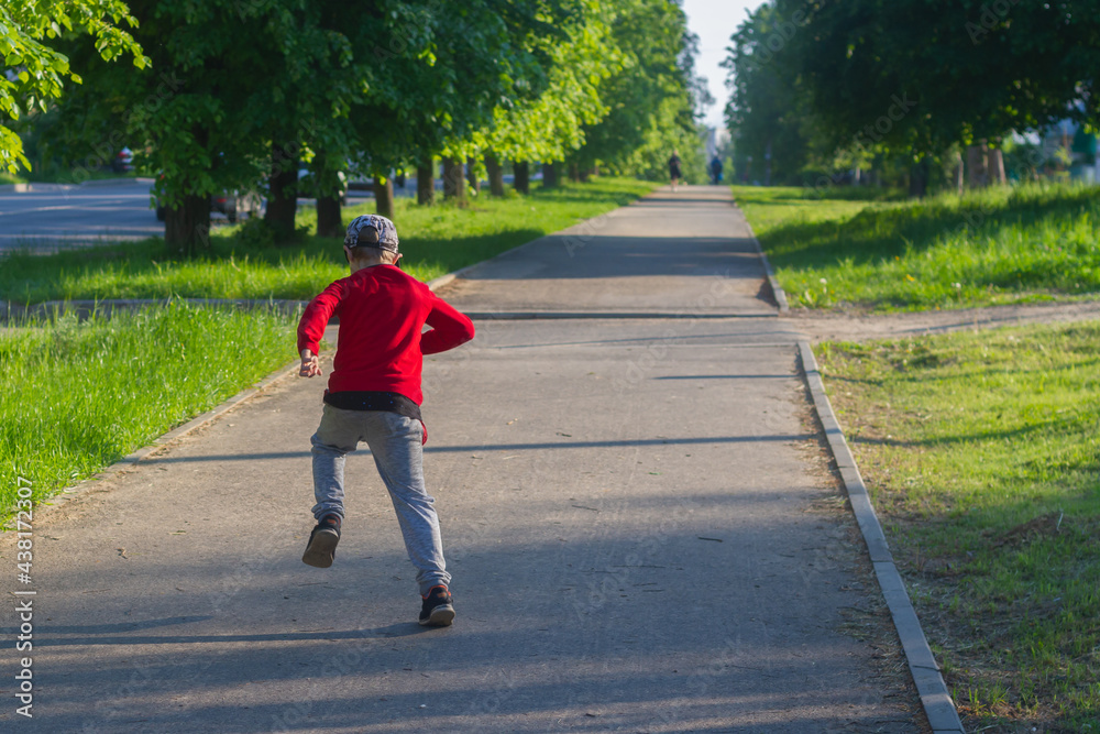 A boy in a red jacket quickly runs along a pedestrian path in the town in summer or spring